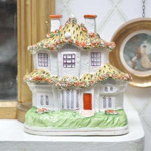 May include: A white ceramic cottage figurine with a thatched roof, orange trim, and a red door. The cottage has multiple windows with purple frames and sits on a green grassy base. Decorative item.