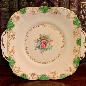 Può includere: Un piatto da portata vintage con bordo smerlato e base color crema. Il piatto presenta un bouquet floreale centrale, rifiniture dorate e dettagli verdi. Ha due manici ed è probabilmente in porcellana.