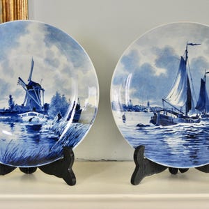Könnte beinhalten: Zwei blau-weiße Zierteller mit Landschaftsszenen. Einer zeigt eine Windmühle, der andere Segelboote auf dem Wasser. Die Teller werden auf schwarzen Holzständern präsentiert und zeigen ein klassisches, maritimes Thema.
