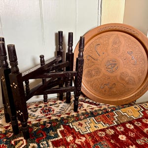 May include: A dark brown wooden folding stand with four legs and small white inlaid accents, positioned next to a large, round, copper-colored metal tray featuring intricate engraved patterns and Arabic calligraphy across its surface.
