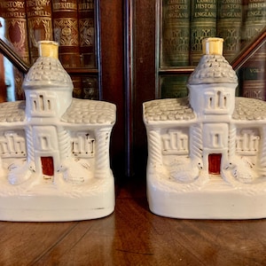 Op de afbeelding: Twee witte keramische kandelaars in de vorm van een huisje met grijze daken en rode deuren. De kandelaars hebben een gedetailleerd ontwerp met ramen en decoratieve elementen. De achtergrond omvat een houten oppervlak en een boekenkast.