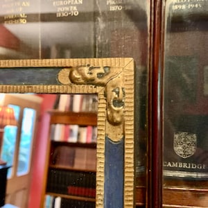 May include: Close-up of a rectangular mirror with a gold and blue frame. The frame has ornate detailing and a crack at the corner. The mirror reflects a room with bookshelves and a lamp. The text "THE ZENITH OF EUROPEAN POWER 1830-70" is visible.