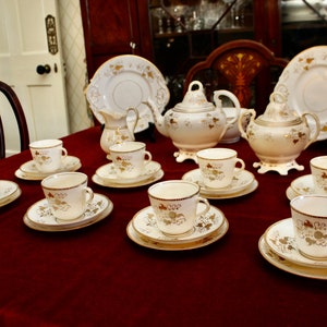May include: A set of white teacups and saucers with gold trim and floral designs. The set includes a teapot, creamer, and sugar bowl. The teacups and saucers are arranged on a red tablecloth.