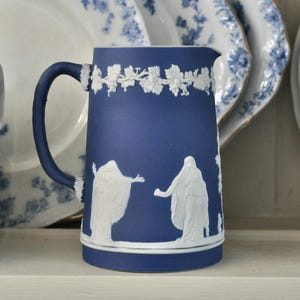 May include: A blue and white ceramic pitcher with a white grapevine design around the top. The pitcher features a white relief design of two figures in flowing robes.