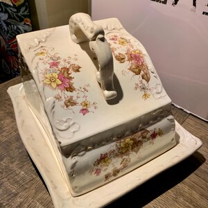 May include: Cream-colored ceramic butter dish with a lid and handle. The dish features a floral pattern in pink, yellow, and brown. The edges have a decorative, embossed design. A vintage-style kitchenware item.