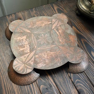 May include: A copper tray with a decorative design of trees, flowers, and geometric patterns. The tray has a scalloped edge and is made of hammered copper.