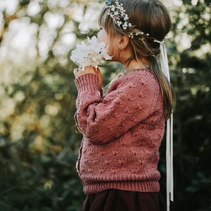 May include: A young girl wearing a pink knitted cardigan with a floral crown and holding a bouquet of white flowers.
