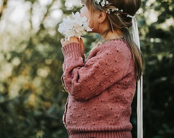 May include: A young girl wearing a pink knitted cardigan with a floral crown and holding a bouquet of white flowers.