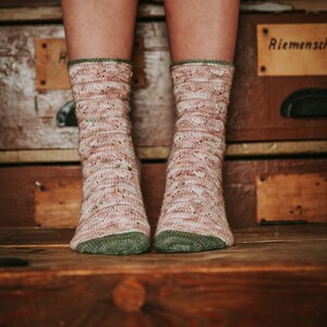 Pode incluir: Um par de meias de malha rosa e verde com um padrão rendado. As meias são usadas por uma pessoa sentada em um baú de madeira com uma etiqueta que diz "Riemensch".