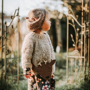 May include: A young child wearing a brown knitted hat and a floral dress with a brown deer-shaped purse. The child is standing in a grassy area with a metal gate in the background.