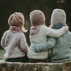 Könnte beinhalten: Drei Kinder in gestrickten Mützen und Pullovern sitzen auf einer Steinmauer. Die Kinder tragen einen hellrosa, einen hellgrauen und einen hellblauen Pullover. Die Kinder sitzen eng beieinander und haben ihre Arme umeinander.