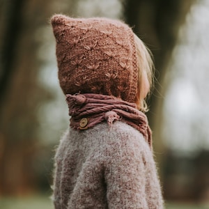 Könnte beinhalten: Ein junges Kind trägt einen rosafarbenen, gestrickten Kapuzenpullover und einen passenden Schal mit einem Holz Knopf. Der Pullover ist flauschig und hat eine lockere Passform.