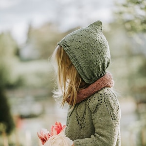 Könnte beinhalten: Ein junges Kind trägt einen grünen gestrickten Kapuzenpullover mit einem rosafarbenen Schal. Es hält einen Strauß aus rosa und weißen Blumen.