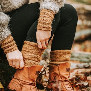 May include: A pair of brown leather boots with laces, worn with brown knitted socks and a grey knitted sweater. The boots are sitting on a bed of fallen autumn leaves.