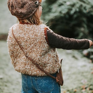 Puede incluir: Un niño lleva una boina de punto marrón, un chaleco texturizado beige y marrón y vaqueros azules con un estampado de lazo. Se lleva un bolso de cuero marrón cruzado. El conjunto es adecuado para otoño o invierno.