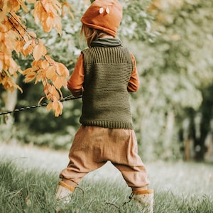 Könnte beinhalten: Ein Kind trägt eine braune Strickweste, eine braune Hose und eine braune Mütze mit Fuchsohren und steht auf einer grasbewachsenen Wiese mit Herbstlaub im Hintergrund.