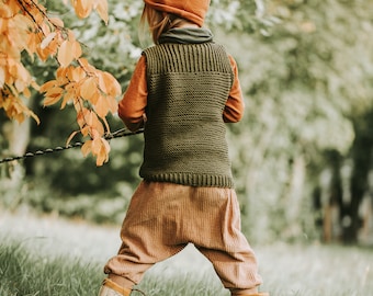 May include: A child wearing a brown knitted vest, brown pants, and a brown hat with fox ears, standing in a grassy field with fall foliage in the background.