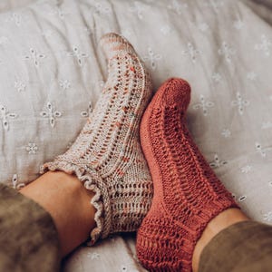 May include: A pair of knitted socks, one beige with colourful flecks and a ruffled edge, the other coral. Both socks feature an openwork design. The socks are handmade and appear to be very comfortable.