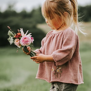 Könnte beinhalten: Ein Kind in einem staubrosa Strickoberteil mit gestickten Gänseblümchen hält einen Blumenstrauß. Der Strauß enthält rosa Dahlien und andere Blüten. Das Kind steht auf einer Wiese. Die Szene hat eine weiche, natürliche Ästhetik.