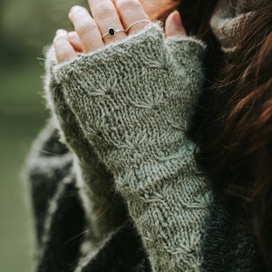 Könnte beinhalten: Nahaufnahme einer Hand, die ein Paar graue, gestrickte Fingerlose Handschuhe trägt. Die Handschuhe haben ein Zopfmuster und sind aus einem weichen, warmen Material gefertigt.