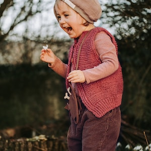 Op de afbeelding: Een jong kind draagt een roze gebreide vest en een bruine corduroy broek, met een bruine leren riem en een houten speelgoed dat aan de riem is bevestigd. Het kind houdt een witte bloem vast en lacht.