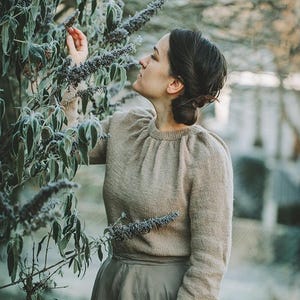 Könnte beinhalten: Eine Frau in einem hellbraunen Pullover und einem langen, fließenden grünen Rock steht in einem Garten und streckt die Hand aus, um einen Zweig mit violetten Blüten zu berühren.