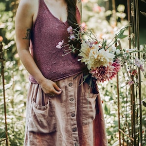 May include: A woman wearing a mauve knit tank top and a button-down skirt in a similar color, holding a bouquet of flowers. The skirt has large pockets. The flowers are a mix of pink, white, and yellow.