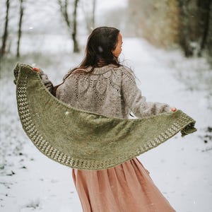 Könnte beinhalten: Eine Frau in einem langen, fließenden rosa Rock und einem braunen Pullover steht in einem verschneiten Wald. Sie hält einen grünen Strick-Schal in ihren Händen.