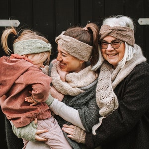 Op de afbeelding: Drie generaties vrouwen dragen gebreide hoofdbanden en sjaals. De jongste wordt vastgehouden door de middelste vrouw, en alle drie lachen.