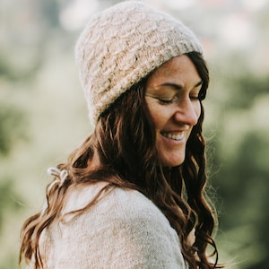 Könnte beinhalten: Eine Frau trägt eine beige Strickmütze und einen Pullover und lächelt mit geschlossenen Augen. Die Mütze hat ein strukturiertes Muster, und der Pullover hat eine weiche, neutrale Farbe. Ihr langes, welliges braunes Haar umrahmt ihr Gesicht, mit einem verschwommenen grünen Hintergrund.