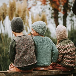Op de afbeelding: Drie kinderen met gebreide mutsen en truien zitten op een houten bank. De kinderen dragen grijze, blauwe en roze mutsen. De truien zijn in groentinten, waarvan één met een geometrisch patroon.