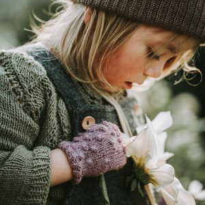 Può includere: Una giovane persona indossa un berretto di lana marrone, un maglione verde e una salopette grigia. Tiene in mano un mazzo di fiori bianchi e indossa dei guanti di lana rosa.