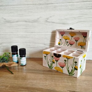 Peut inclure: Une boîte en bois blanc avec un motif floral sur le couvercle et les côtés. La boîte a un couvercle à charnières et est divisée en compartiments. Deux petites bouteilles avec des bouchons noirs sont devant la boîte.