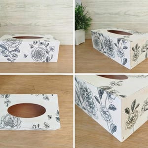 May include: A white rectangular tissue box featuring a black floral pattern. The box has an oval aperture on top and is adorned with roses, leaves, and butterflies. The box is made of wood.