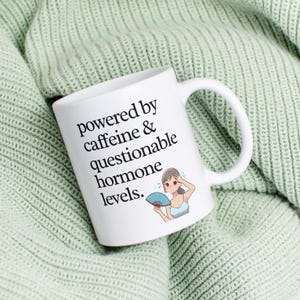 May include: White ceramic mug with the text "powered by caffeine & questionable hormone levels." A cartoon woman with a fan is also printed on the mug. The mug is resting on a light green knitted blanket.