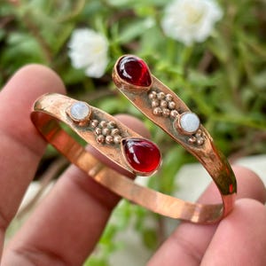Peut inclure: Un bracelet manchette en cuivre au design ouvert, orné de deux pierres rouges en forme de goutte et de deux petites pierres blanches rondes. La surface métallique présente une finition texturée avec des grappes perlées autour des pierres.