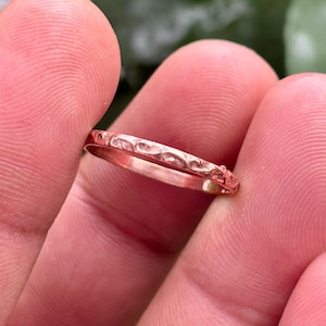 Puede incluir: Primer plano de un anillo de oro rosa con un diseño texturizado y estampado. El anillo se sostiene entre dos dedos, mostrando sus delicados detalles. La superficie del anillo tiene un patrón repetitivo de pequeñas formas curvas.