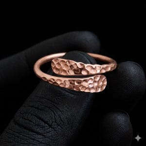 May include: A copper-coloured open ring featuring a hammered, textured surface. The band has a bypass design where the two ends overlap slightly, showcasing a consistent dimpled pattern across the entire outer circumference of the metal ring.
