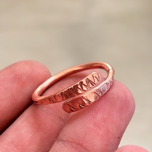 May include: A copper-colored bypass ring featuring a textured surface with repeating diagonal etched marks. The band is open-ended, allowing for adjustment, and displays a metallic finish with a warm, reddish-brown hue throughout the piece.