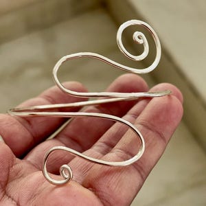 May include: A silver arm cuff with a swirling design. The cuff features two curved sections that wrap around the arm, with spiral accents at the ends. The metal has a polished finish, reflecting light. This piece of jewelry is designed to be worn on the upper arm.