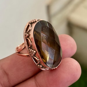 May include: A copper-colored ring featuring an oval, faceted, smoky quartz gemstone. The ring has an ornate, decorative setting with floral and vine-like details. The gemstone is a deep brown color and catches the light.