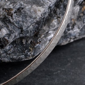 Op de afbeelding: Een zilveren gehammerde armband met een gladde, gepolijste afwerking. De armband ligt op een grijze rots.
