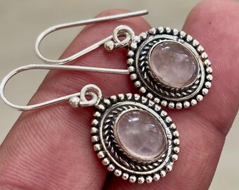 May include: A pair of silver-toned earrings with oval, light pink gemstones. The earrings have a decorative border with a twisted rope design and small beaded accents. The ear wires are curved.