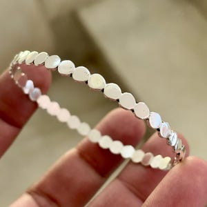 May include: A silver bracelet with a repeating pattern of small, flat, circular shapes. The bracelet is held in a hand, and the design is visible against a blurred background. The bracelet is a piece of jewelry.