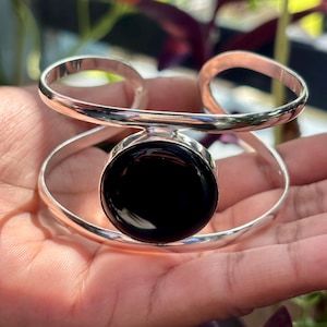May include: A silver-toned cuff bracelet with a large, round, black stone at its center. The bracelet has a modern design with two curved bands that wrap around the wrist. The polished metal reflects light, creating a bright, eye-catching accessory.