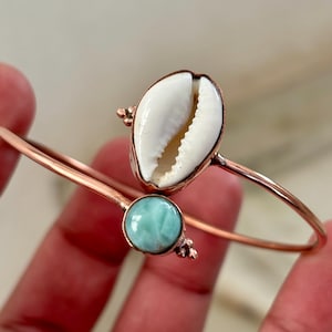 Könnte beinhalten: Ein kupferfarbenes, offenes Armband mit einer weißen Kaurimuschel und einem runden, glatten, hellblauen Edelstein. Das Metallband ist dünn und poliert, mit kleinen dekorativen Metallakzenten, die die Muschel und den Stein an den Enden flankieren.