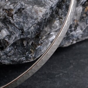 Op de afbeelding: Een zilveren gehammerde armband met een gladde, gepolijste afwerking. De armband ligt op een grijze rots.