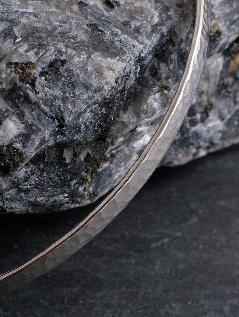 Op de afbeelding: Een zilveren gehammerde armband met een gladde, gepolijste afwerking. De armband ligt op een grijze rots.