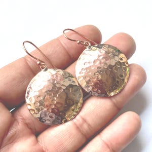 May include: A pair of copper-colored, hammered-metal disc earrings. Each earring features a circular design with a textured, dimpled surface and a simple hook for the ear. The earrings are displayed against a light background.