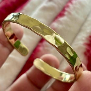 May include: A gold-coloured cuff bracelet with a hammered texture. The open-ended design allows for easy wear. The bracelet is shown against a soft, patterned background.
